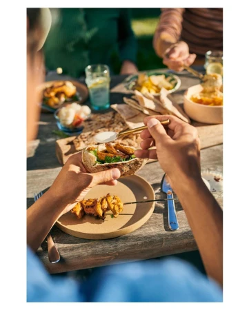 Person adding sauce to food on pita bread over a wooden table with other small bowls, glasses, and utensils outdoors in daylight.
