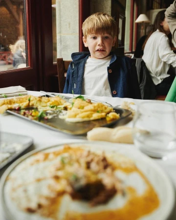 Young boy at restaurant table with leftover food, wearing a denim jacket, sitting at a table with plates and glasses.