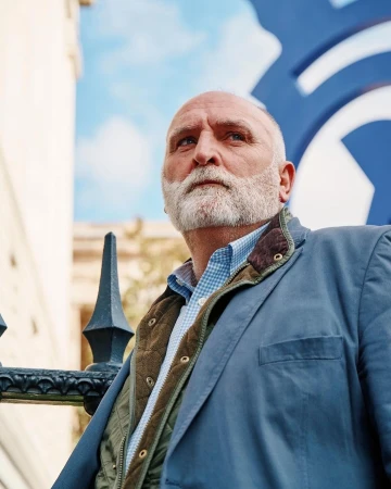Low-angle portrait of an older man with a white beard wearing a blue jacket and plaid shirt outdoors in London by Peden+Munk.