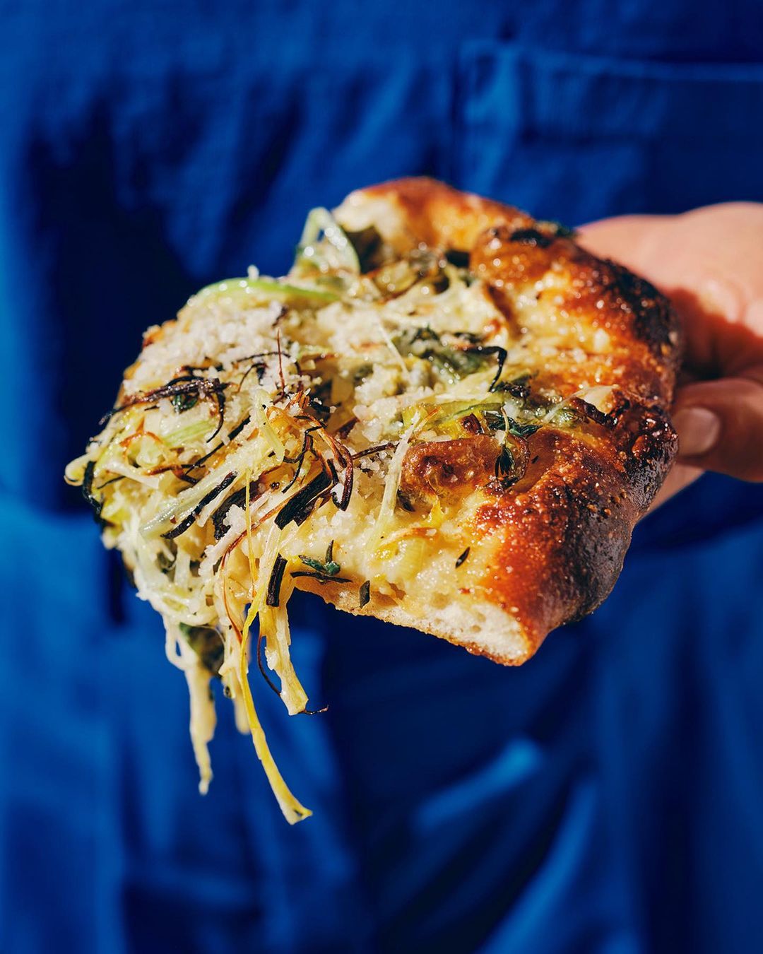 Close-up shot of a triangular slice of pizza with melted cheese and green vegetables, held by a human hand against a blue background.