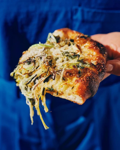 Close-up shot of a triangular slice of pizza with melted cheese and green vegetables, held by a human hand against a blue background.