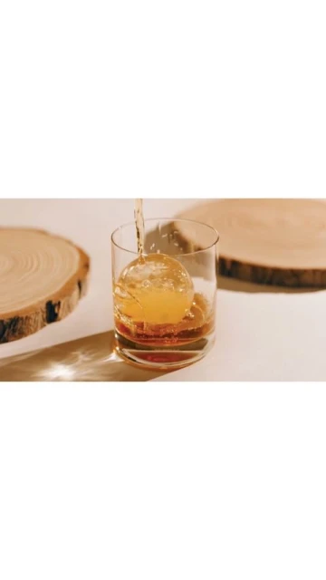 Amber liquor being poured into a clear glass with an ice ball, set on a white surface with wood slices.
