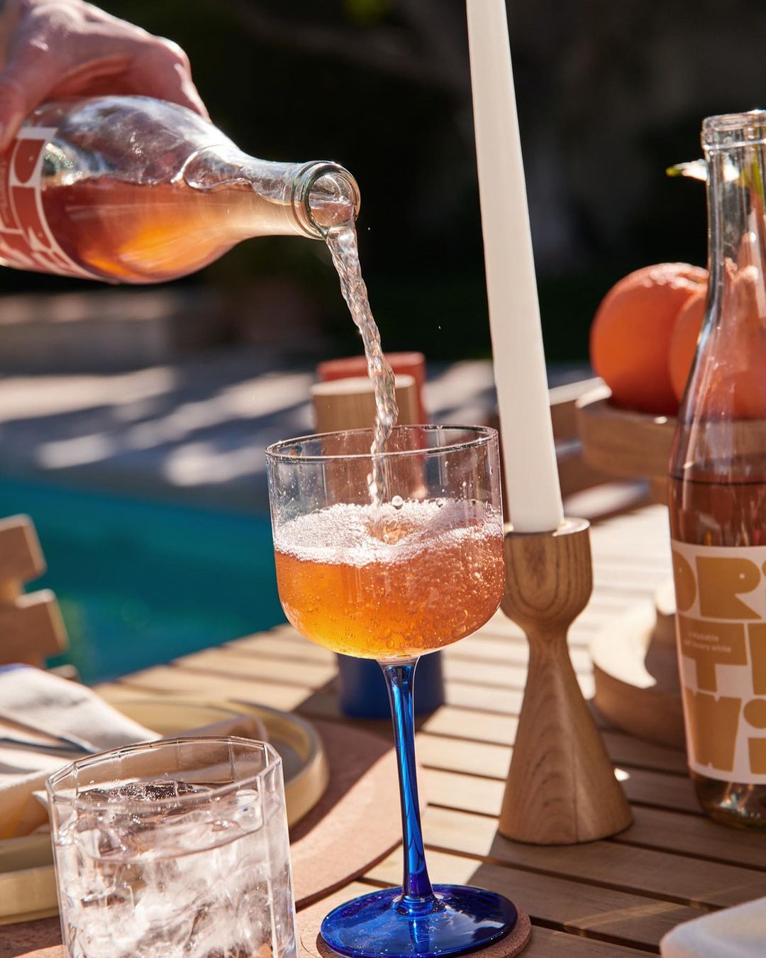 Refreshing drink being poured into a stemmed glass on a wooden table outdoors, with oranges and a candle nearby.