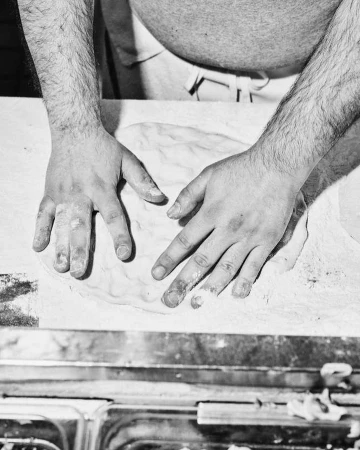 Overhead monochrome shot of a baker's hands kneading dough on a floured surface, with a glimpse of stainless steel equipment below.