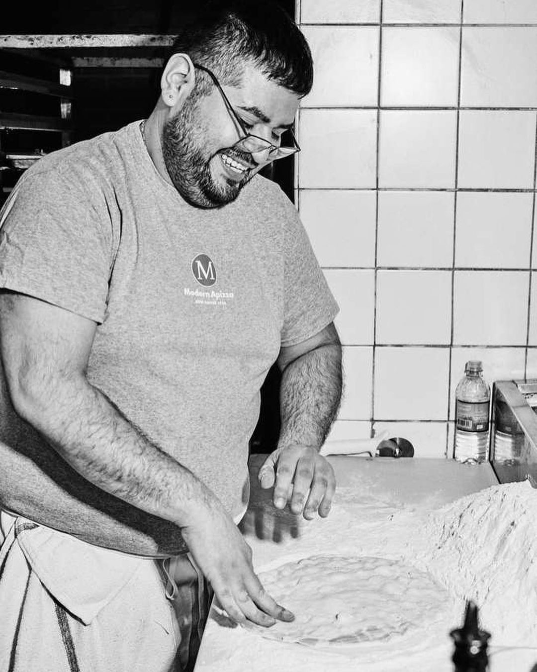 A smiling adult male wearing glasses prepares dough in a kitchen, with a bottle and white tiled wall in the background, in black and white.