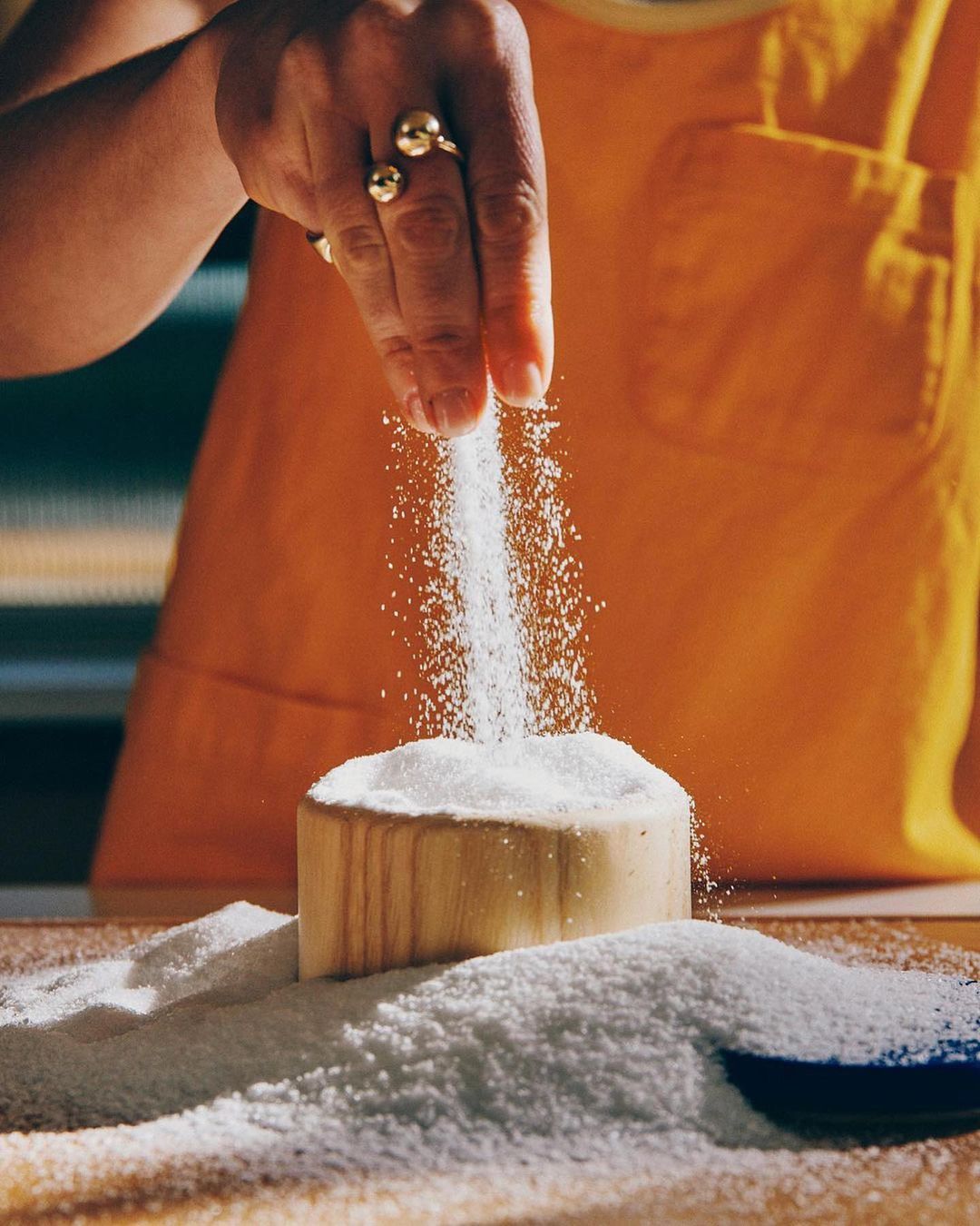 Hand sprinkles white granules from above onto a wooden bowl filled with the same. Granules are also scattered on a wooden surface.