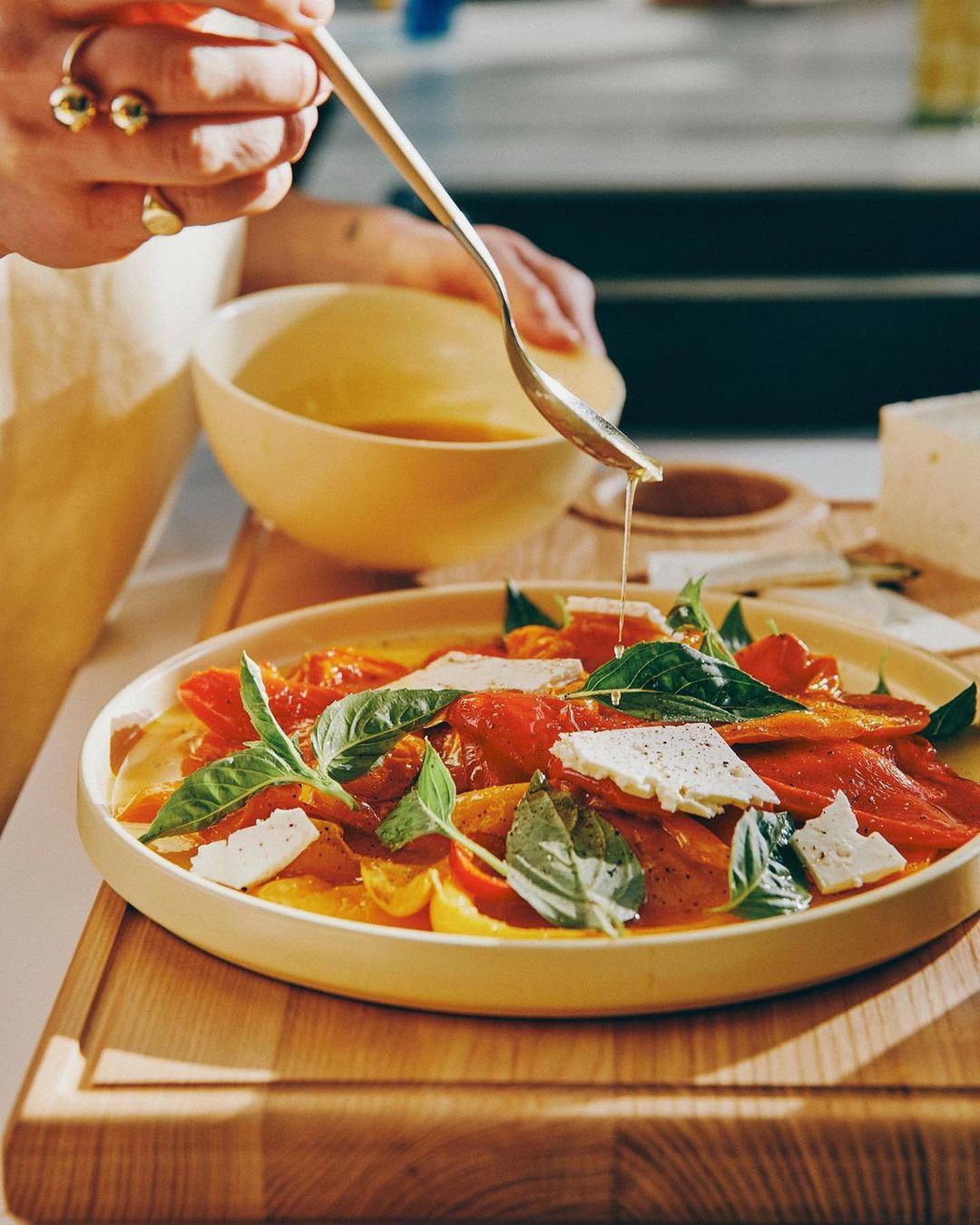 Overhead shot of a person dressing a colorful salad of peppers, feta, and basil with a golden spoon, set on a wooden board.