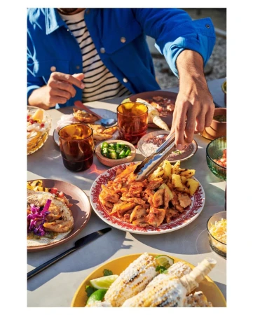 Overhead shot of a person reaching for food with tongs at an outdoor table laden with tacos, corn, and other small dishes.