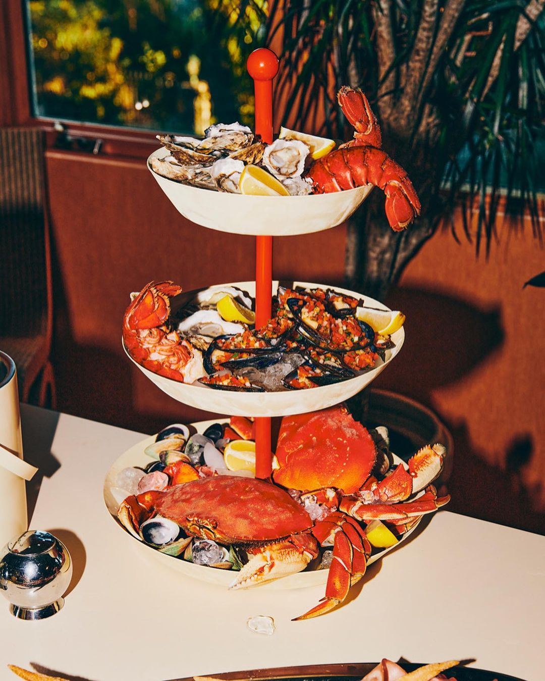 Three-tiered seafood tower with oysters, lobster, and crab on a table with a muted color palette, lit by natural light.