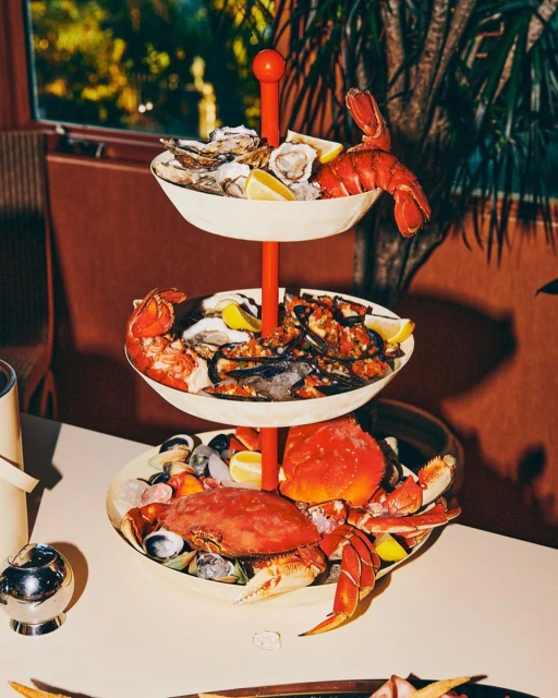 Three-tiered seafood tower with oysters, lobster, and crab on a table with a muted color palette, lit by natural light.
