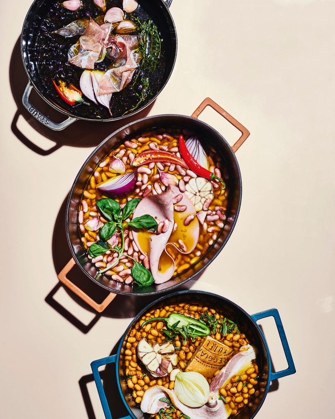 Overhead shot of three Staub pots filled with ingredients like beans, garlic, onions, meat, and herbs against a neutral backdrop.