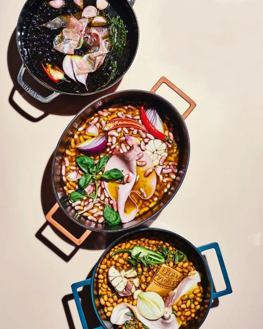 Overhead shot of three Staub pots filled with ingredients like beans, garlic, onions, meat, and herbs against a neutral backdrop.