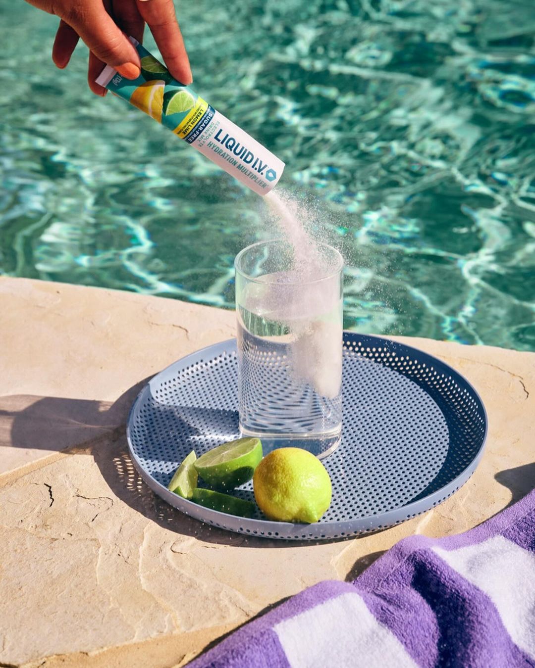 Powder being poured from Liquid I.V. packet into glass of water on tray with lime and lemon, poolside on sunny day.
