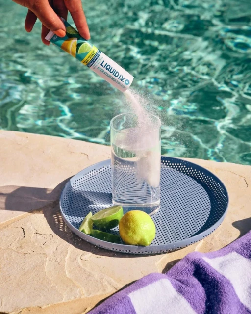 Powder being poured from Liquid I.V. packet into glass of water on tray with lime and lemon, poolside on sunny day.