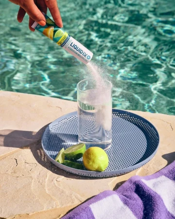 Powder being poured from Liquid I.V. packet into glass of water on tray with lime and lemon, poolside on sunny day.