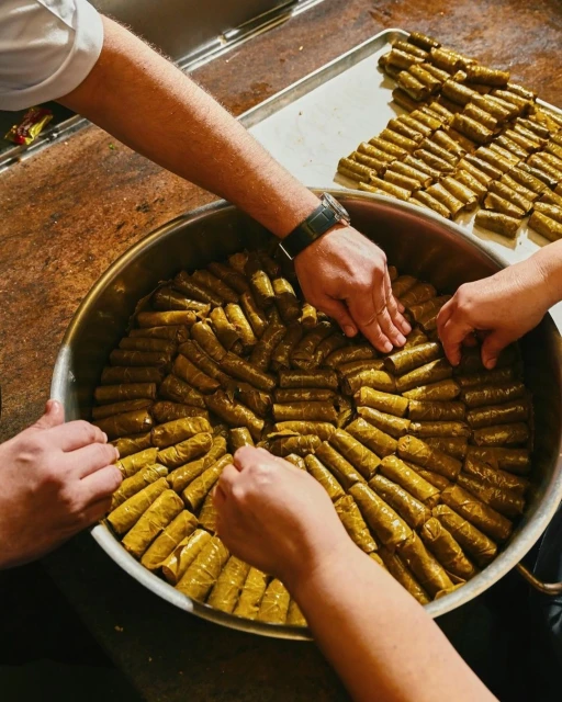 Overhead shot of several people's hands arranging stuffed grape leaves in a large, round metal serving tray, with a platter of rolls behind.