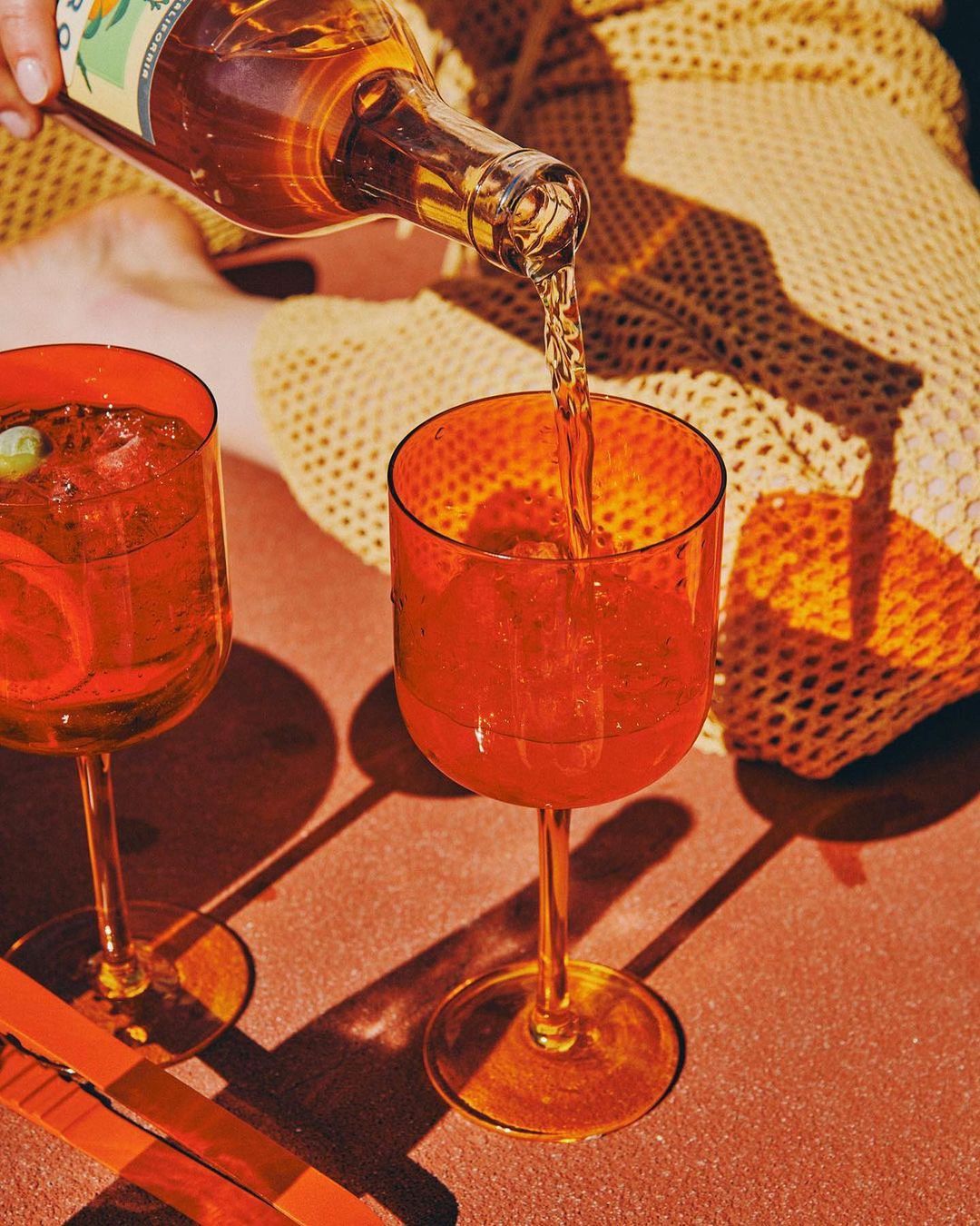 Close-up of amber liquid being poured into a vibrant orange glass; a second cocktail glass is visible alongside a patterned blanket.