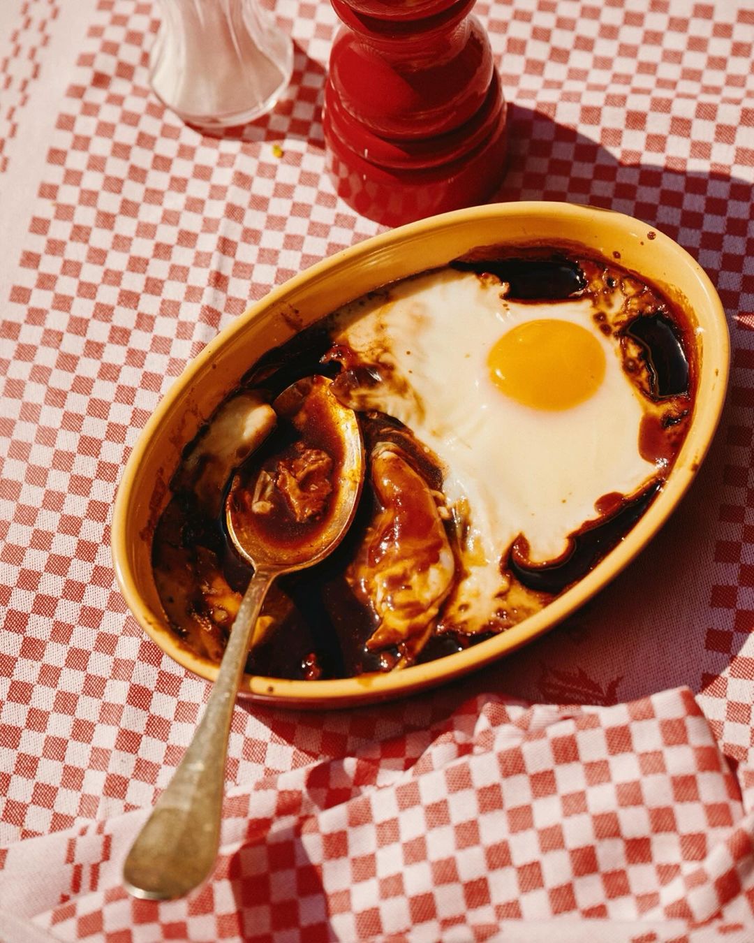 Overhead shot of a cooked egg on top of a dish with brown gravy, served on a red and white checkered tablecloth.
