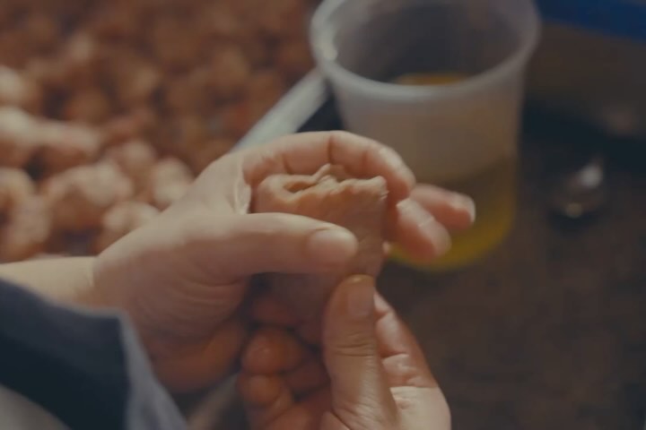 Close-up of two hands shaping food dough, with blurred dough and a bowl of yellow liquid in the background, indoor setting.
