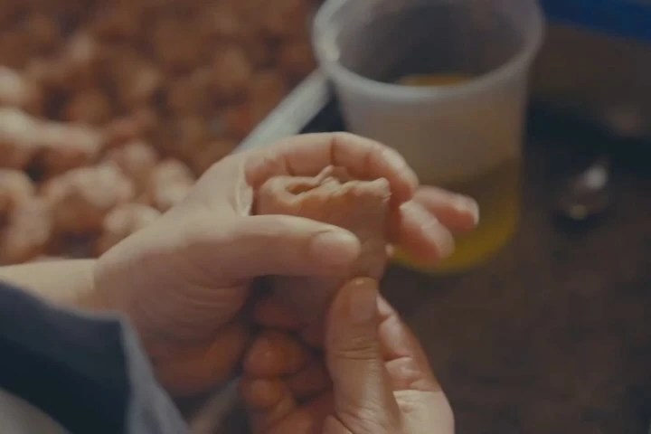 Close-up of two hands shaping food dough, with blurred dough and a bowl of yellow liquid in the background, indoor setting.