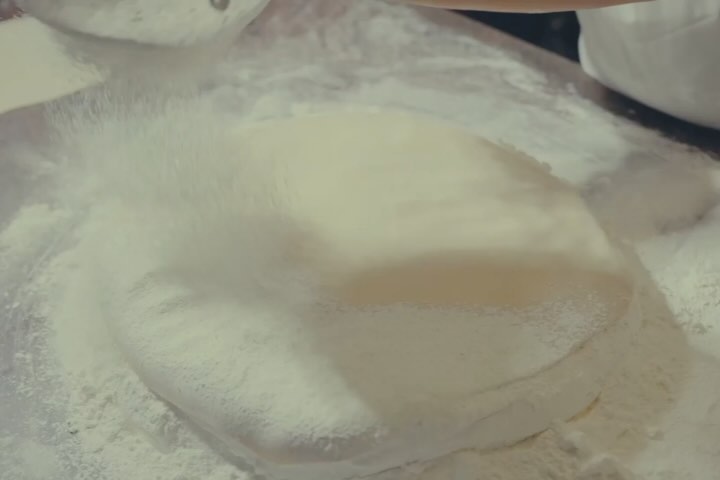 Close-up of dough being dusted with flour, creating a cloudy effect, on a lightly-colored surface, suggesting a baking or cooking preparation scene.