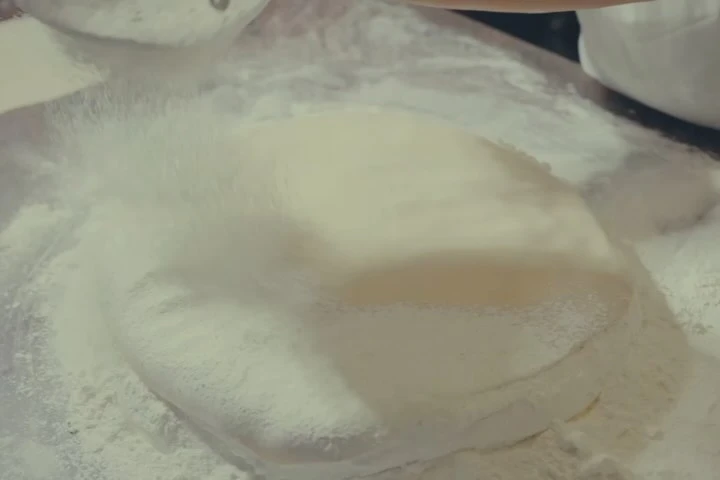 Close-up of dough being dusted with flour, creating a cloudy effect, on a lightly-colored surface, suggesting a baking or cooking preparation scene.