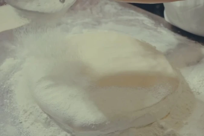 Close-up of dough being dusted with flour, creating a cloudy effect, on a lightly-colored surface, suggesting a baking or cooking preparation scene.