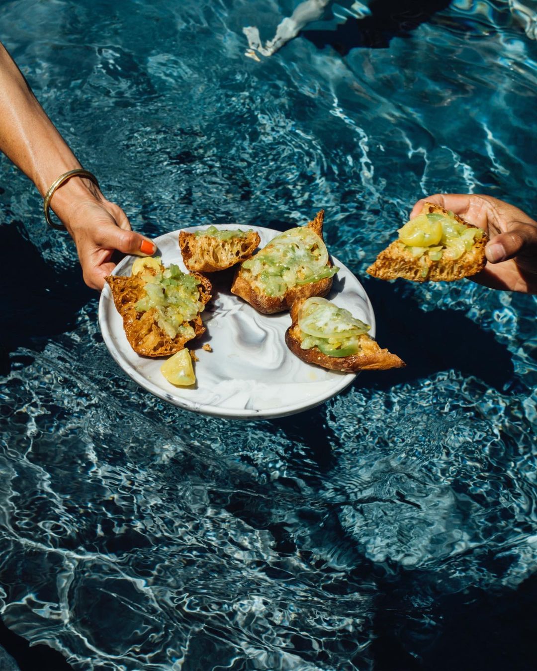 Overhead shot in pool: hands hold plate with bread and dipping sauce near water with rippled textures and light patterns.