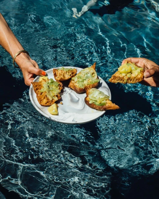 Overhead shot in pool: hands hold plate with bread and dipping sauce near water with rippled textures and light patterns.