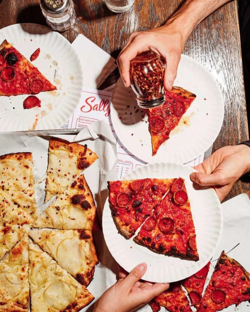 Overhead shot of hands holding plates of pepperoni pizza and cheese pizza on a wooden table with a jar of chili flakes.