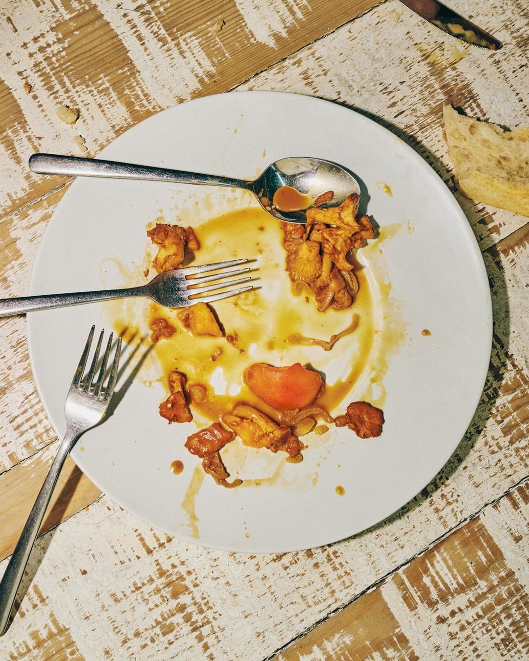Overhead shot of a white plate with the remnants of a meal, featuring chanterelle mushrooms, tomato, fork and spoon on a rustic table.