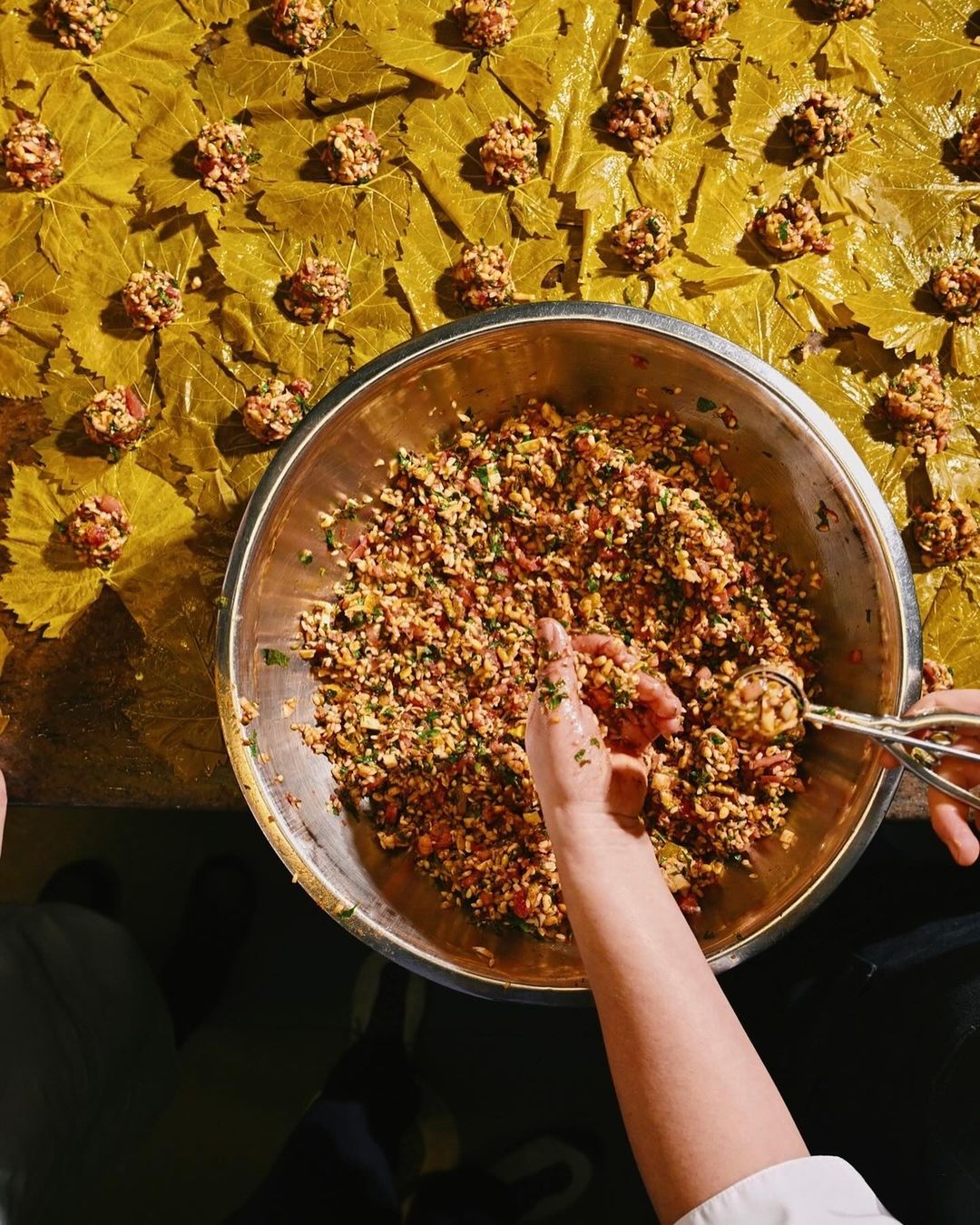 Overhead shot showcases a person using a scoop to fill grape leaves on a table with food preparation. Indoors with diffused lighting.