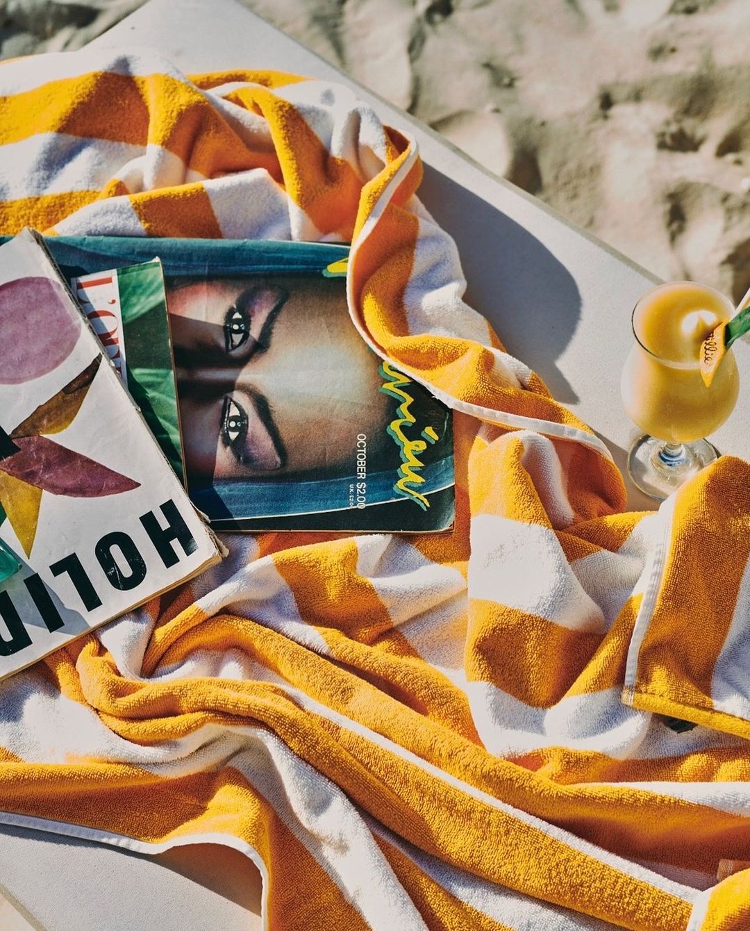 Beach scene with a striped towel, fashion magazines, and a tropical drink creates a relaxed, summery vibe for travel or leisure content.