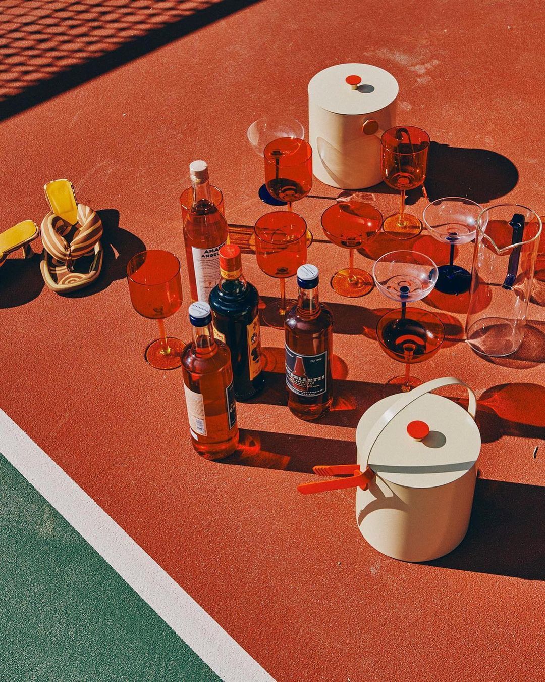 Still life of beverage bottles and glasses filled with colorful liquids, arranged on a red and green surface, with soft light and shadows.