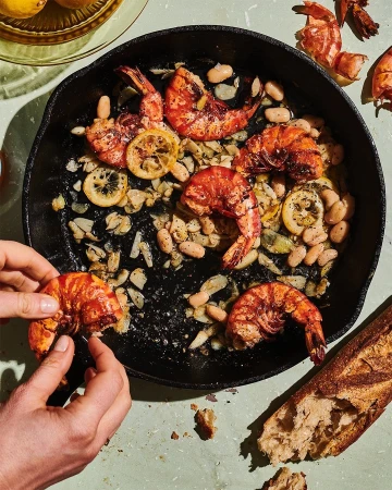 Overhead shot of hands preparing shrimp and white bean skillet with sliced lemons, served with baguette on a light green surface.