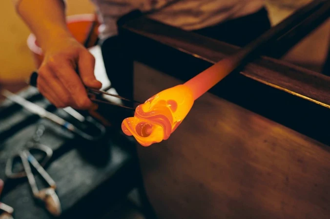 Close-up of a glassblower shaping molten glass with metal tools, creating an orange glass art piece indoors.
