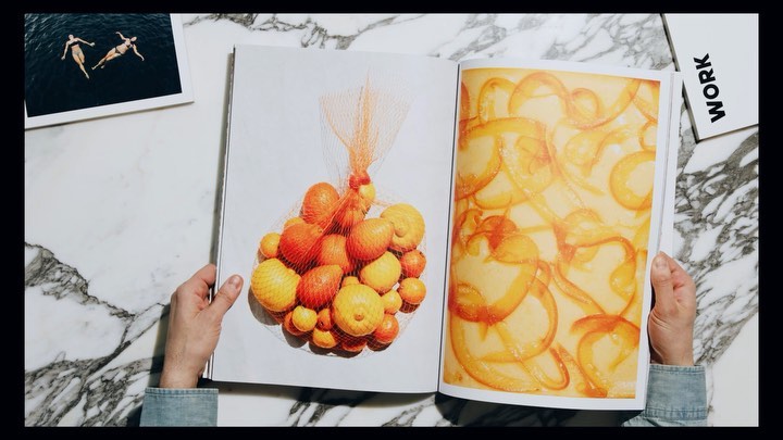Open magazine pages show oranges and abstract orange shapes on white marble table with a photo of two women swimming nearby.
