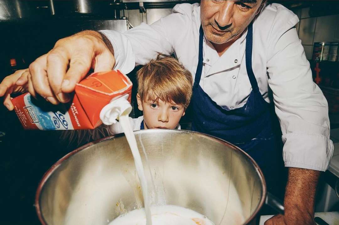 Chef pours milk into a mixing bowl, with a child observing in a commercial kitchen. Food preparation, culinary arts, collaboration.