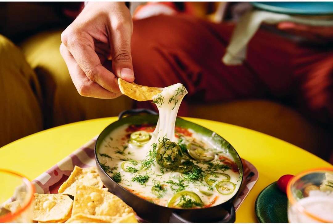 Close-up of a hand dipping a tortilla chip into a dish of melted cheese with jalapeños and dill on a yellow table.
