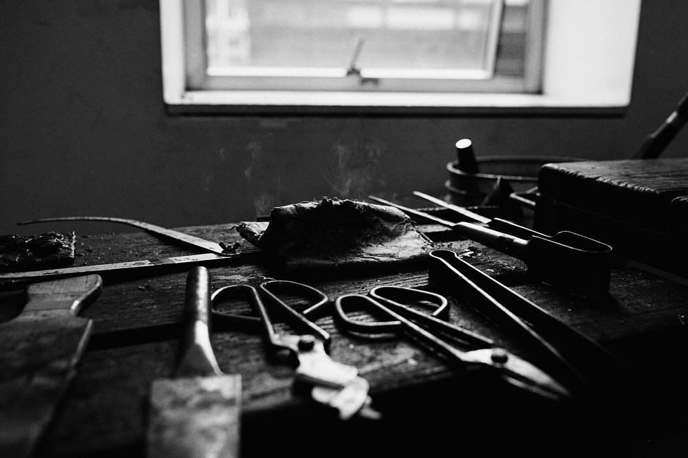 Black and white photo of a workbench with assorted tools including scissors, tongs, knives, and a hammer under soft window light.