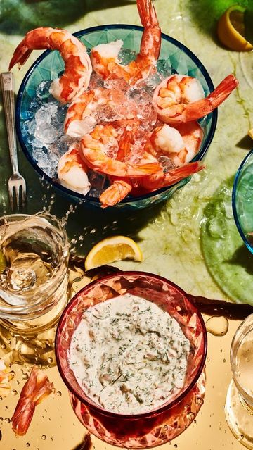 Overhead shot of shrimp cocktail in blue bowl with cocktail sauce in pink bowl, lemon slice, and water droplets on table.