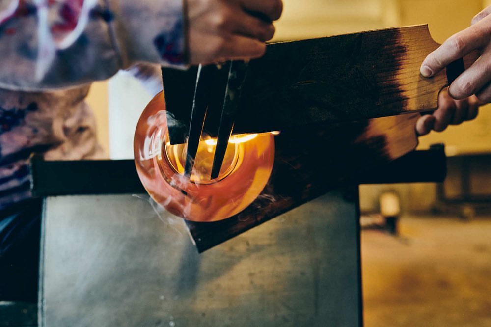 Glass blowing process with molten glass being shaped by a glassblower using hand tools in a dimly lit studio environment.