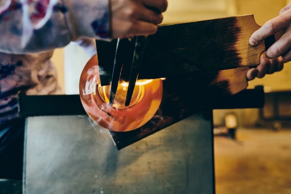 Glass blowing process with molten glass being shaped by a glassblower using hand tools in a dimly lit studio environment.
