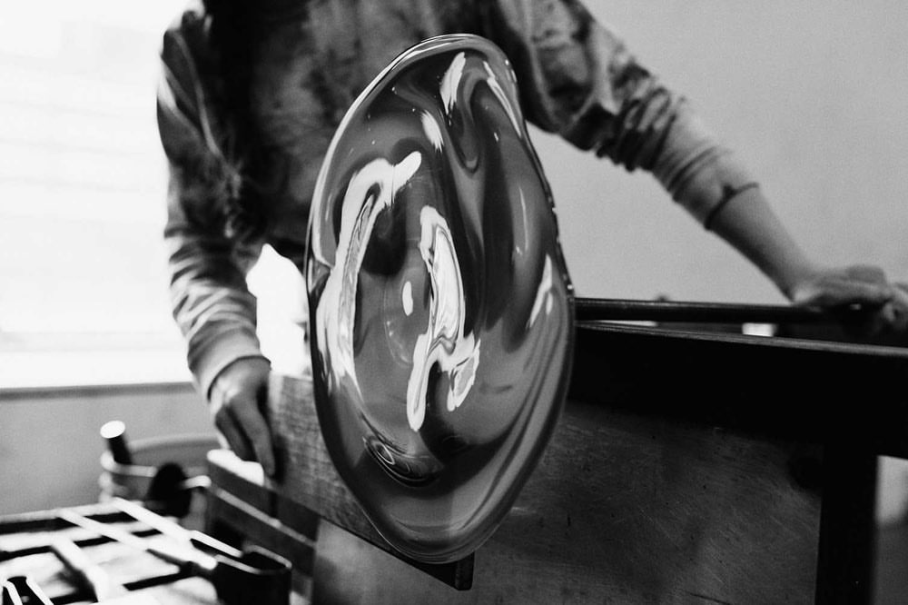 Black and white image of a glassblower shaping molten glass with hands and tools in a studio setting, creating a unique art piece.