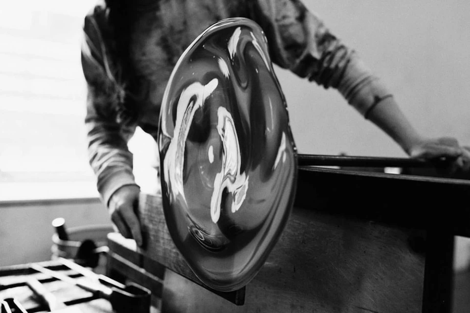 Black and white image of a glassblower shaping molten glass with hands and tools in a studio setting, creating a unique art piece.