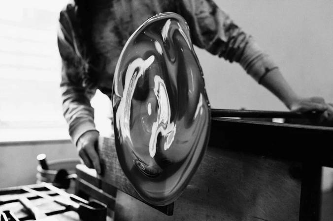 Black and white image of a glassblower shaping molten glass with hands and tools in a studio setting, creating a unique art piece.