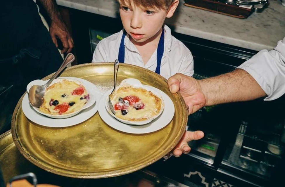 Young boy looking at two desserts on a gold tray held by a server in a restaurant, featuring fresh fruit and powdered sugar.