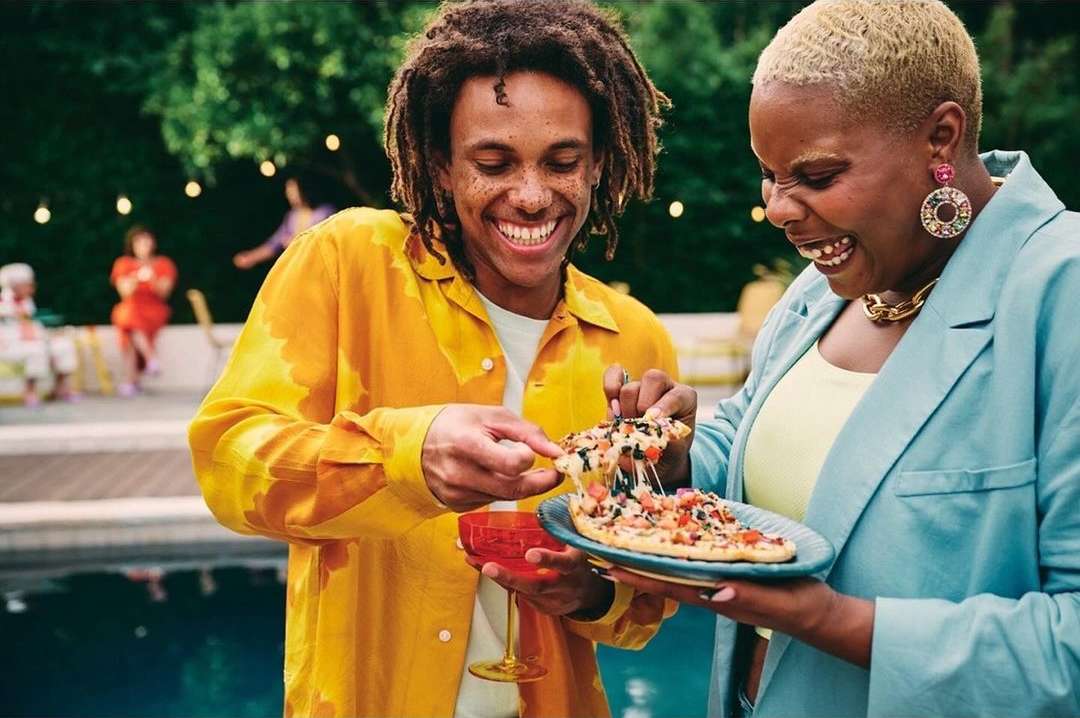 Two adults smiling outdoors while sharing a slice of pizza with melting cheese at an outdoor gathering by a pool surrounded by trees.