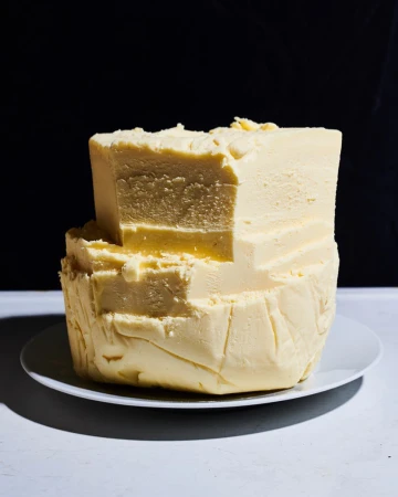 Stack of butter on a white plate against a dark background, showcasing its texture and rich color in a studio food photography shot.