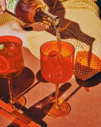 Amber liquid being poured from a glass bottle into a textured orange wine glass on a sunlit tabletop.